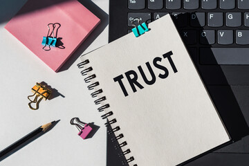 Trust word printed on notepad lying on laptop next to pink sticknotes, paper clips and black pencil.