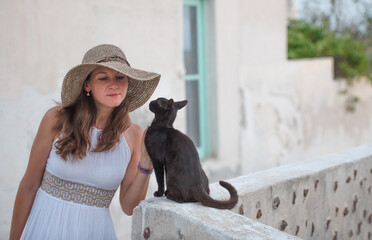 Young beautiful woman in white long dress and sun hat touching a black street cat in Santorini...