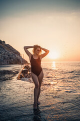 Fototapeta na wymiar Beautiful young woman in a black swimsuit alone on the beach by the sea at sunset. An adult slender girl resting on the ocean shore at sunset. Summer vacation concept. Selective focus