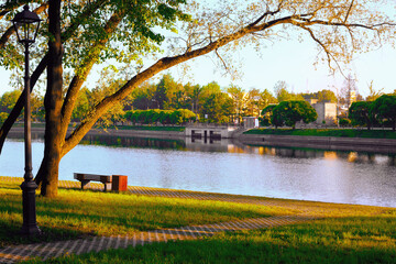 Obraz premium Morning beautiful calm landscape: a street lamp, trees, green grass, a bench and an embankment on the river bank, illuminated by the rising rays of the sun.