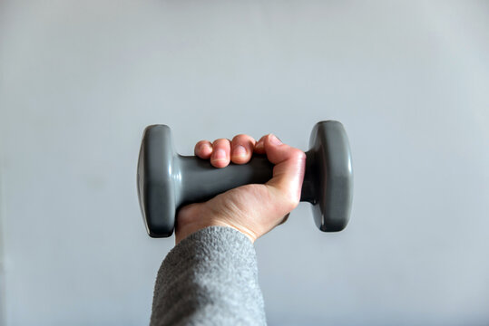 Detalle del brazo de una mujer con una sudadera gris agarrando una mancuerna peque&ntilde;a sobre fondo blanco.