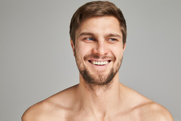 Fototapeta premium Portrait of a smiling young man with bare swimmers shoulders on a gray background, powerful, beard, charismatic, adult, brutal, athletic, edited photo, bright smile, white teeth smile, look in camera