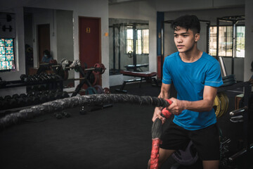 A fit young asian man working out vigorously with battle ropes. Alternating single arm waves. Upper body workout, conditioning and cardio at the gym.