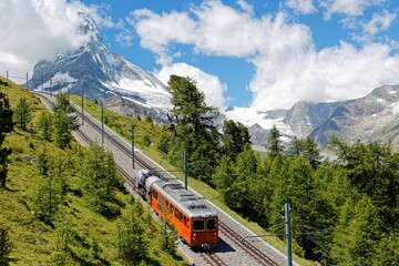 A cogwheel train traveling on Gornergrat railway through a forest by the mountainside with majestic Matterhorn mountain peak in background on a cloudy sunny summer day, in Zermatt, Valais, Switzerland