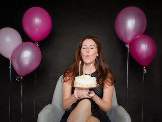 Pretty woman blowing candles out on birthday cake with balloons.
