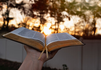 Close up woman outstretching hand with open Bible near bright sunset in the background. copy space.