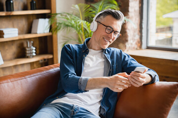 Smiling caucasian mature middle-aged man freelancer relaxing resting listening to the music radio podcast playlist choosing soundtrack on cellphone at home office