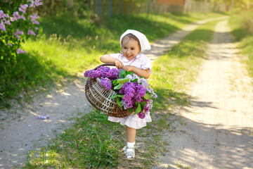 A village girl in a white dress and a kerchief carries a basket filled with spring blooming flowers and violet lilac along a country path. Funny happy pretty little child collects flowers in a bouquet