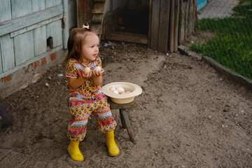 Baby is sitting on a bench by the chicken coop and holding fresh eggs in her hands. Next to the girl is a hat filled with chicken eggs from a laying hen. The child looks towards copy space © Maryna