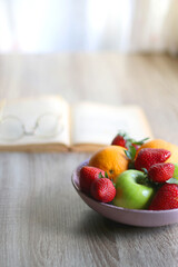 Bowl of apples, oranges and strawberries, open book and reading glasses on the table. Selective focus.