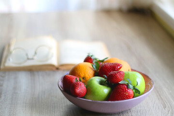 Bowl of apples, oranges and strawberries, open book and reading glasses on the table. Selective focus.