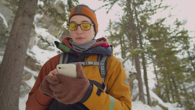 Arc waist up shot of female tourist with backpack checking map on smartphone while hiking in mountain forest in winter