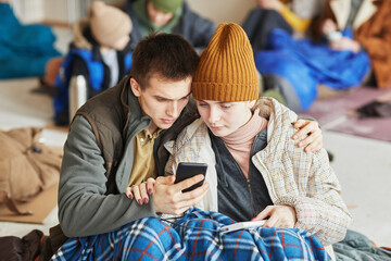 Front view portrait of young Caucasian couple hiding in refugee shelter covered with blanket on...