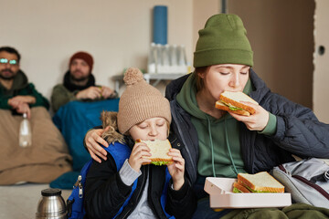 Portrait of Caucasian young mother and son eating sandwiches in refugee shelter during war or...