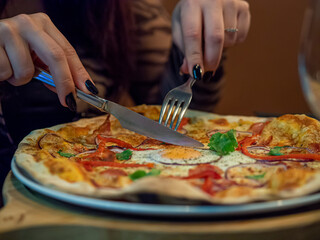 Woman's hand holding fork and knife and cutting a delicious thin crust pizza. Close up of pizza plate with fry egg, cheese and vegetables. Dinner with friends in an Italian restaurant.