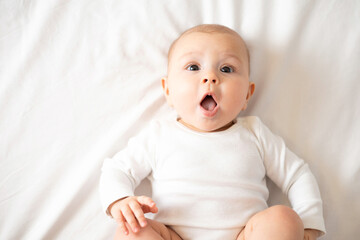 Portrait of a cute baby girl of 5 months in a white bodysuit on a white bed in the bedroom. Space for text. Child lifestyle