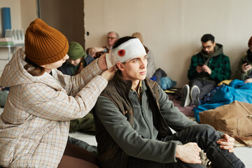 Portrait of young woman bandaging head of wounded young man in refugee shelter, copy space