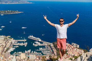 Fototapeta na wymiar A handsome young man stands and jubilantly with his hands raised on a mountain with the name Of a Head of dog, the Principality of Monaco in the background in clear sunny weather