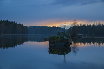 Early sunrise on a small calm Finnish lake with a large stone and trees in the middle, blue sky above blue water