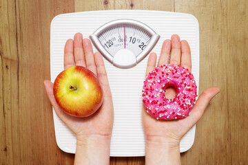 Diet concept. Female hands hold sweet donut and red apple against the background of scales, choice...