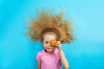 A beautiful white girl in a pink T-shirt lies with her blond hair loose in a semicircle on a blue background in the center, closes her left eye with round brown oatmeal cookies and smiles, top view