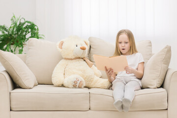 Photo of plush teddy bear and little girl holding a book on sofa.