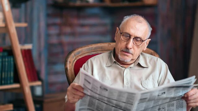 Focused elderly man reading paper newspaper text relaxing in armchair at rustic home interior