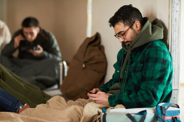 Side view portrait of young man hiding in shelter during war or crisis and holding smartphone - obrazy, fototapety, plakaty