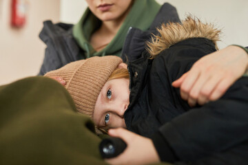 Portrait of blonde Caucasian child lying in mothers lap at refugee shelter and looking at camera...