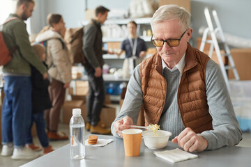 Portrait of Caucasian senior man eating simple meal at soup kitchen in help center for refugees, copy space
