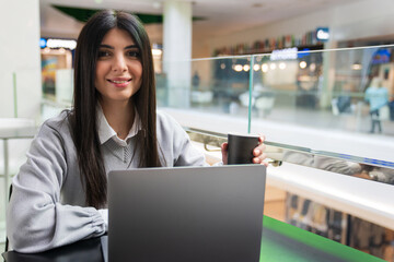 A young woman drinks coffee and works at a laptop in a shopping center cafe