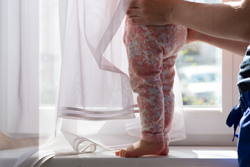 baby girl stands on the windowsill with curtain, baby feet, mother holds child at sunny day, home window background © skypictom