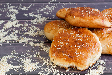 Homemade cakes with yeast, fragrant golden buns sprinkled with sesame seeds, a lot of white sesame seeds scattered around, crispy bread on the table