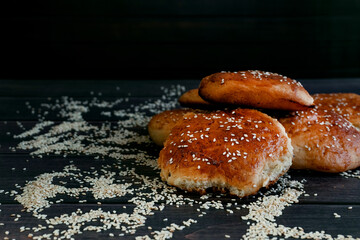 Homemade cakes with yeast, fragrant golden buns sprinkled with sesame seeds, a lot of white sesame seeds scattered around, crispy bread on the table
