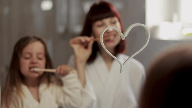 Close-up of beautiful young mother and her cute daughter in white bathrobe brushing teeth standing in front of a mirror with drawing heart in the bathroom.