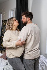 Fototapeta premium Happy family concept. Husband hug belly pregnant wife standing indoor living room near sofa
