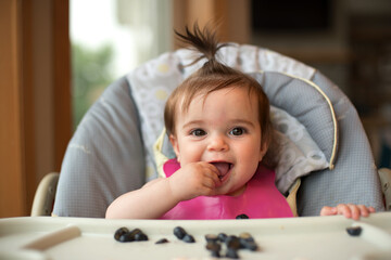 A smiling baby with a ponytail sits in a highchair and eats blueberries