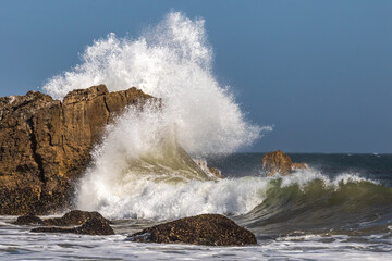 Large wave breaking against giant rock formation offshore near Malibu, California. Spray flying high into the air. Sky, Pacific Ocean in the distance. 

