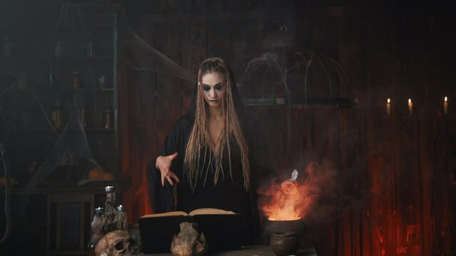 Halloween, witch reading magic book use cauldron prepare poison or love potion. Black magic occult female wizard in dark room cage spider web human skull Necromancer. voodoo. Handheld