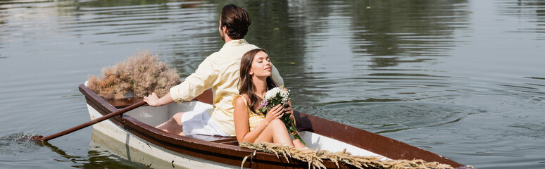 young woman holding flowers and leaning on back of boyfriend during romantic boat trip, banner.
