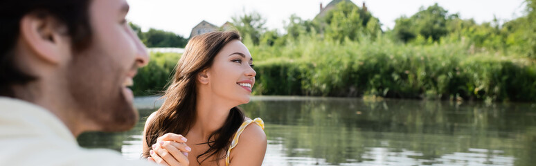 happy woman smiling and looking away near cheerful and blurred boyfriend, banner.