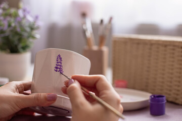 Close-up of female hands paint with a brush a sprig of a purple lavender , on a white ceramic pot.