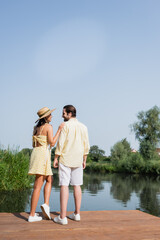 Fototapeta na wymiar full length of happy young couple in summer clothes holding hands and standing on pier near lake.