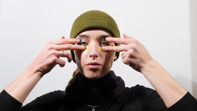 A girl in a green hat on a white background, paints a camouflage makeup with her fingers in the colors of the Ukrainian flag, yellow-blue