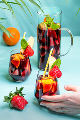 A female hand holding a glass with traditional Spanish Sangria cocktail decorated with pieces of fresh strawberry, pinch of mint and eco-paper straw.