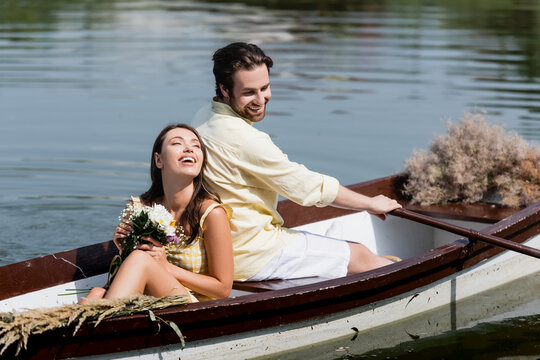 happy young woman holding flowers and leaning on back of boyfriend during romantic boat trip.