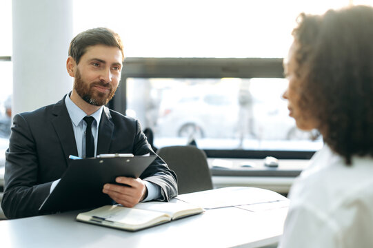 Interview. Young adult caucasian male, hiring manager, sits at a desk in a modern office, conducts an interview with an African American girl, looks through a resume, talks about working conditions