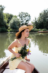 happy young woman in straw hat and dress holding flowers while having boat ride on lake.