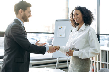 Business partners, man and woman of different nationalities, stand in modern office, hold business negotiations, made a good deal, agreed on cooperation, greet each other shaking hands, smiling