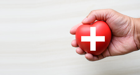 A red cross is a symbol of medical health. Hand holding a heart and on a wooden desk. Copy space...
